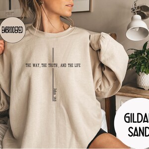 May include: Sand-colored embroidered sweatshirt with the text "THE WAY, THE TRUTH, AND THE LIFE" and "John 14:6" in black. The sweatshirt has a crew neck and long sleeves. The brand name "GILDAN SAND" is also visible.