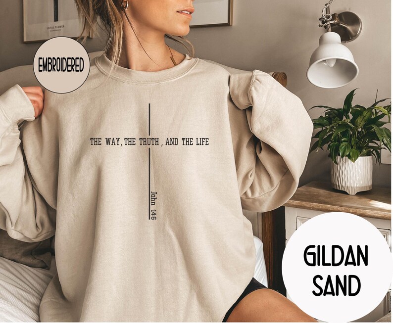 May include: Sand-colored embroidered sweatshirt with the text "THE WAY, THE TRUTH, AND THE LIFE" and "John 14:6" in black. The sweatshirt has a crew neck and long sleeves. The brand name "GILDAN SAND" is also visible.