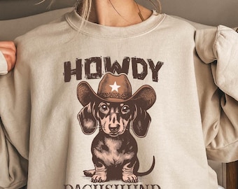 May include: Beige sweatshirt featuring a graphic of a dachshund wearing a cowboy hat with a star. The word "HOWDY" is above the dog, and "DACHSHUND" is below. The sweatshirt has long sleeves.