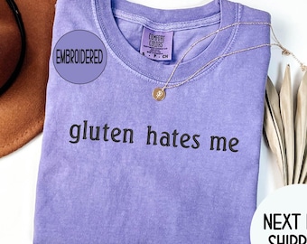 May include: A light purple t-shirt with the embroidered text "gluten hates me" in black. The shirt has a Comfort Colors tag and a gold necklace. A brown hat is visible in the upper left corner.