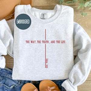 May include: Light gray embroidered sweatshirt with the text "THE WAY, THE TRUTH, AND THE LIFE" in red, with "John 14:6" below. The sweatshirt has a crew neck and long sleeves. A circular patch that says "EMBROIDERED" is on the upper left.