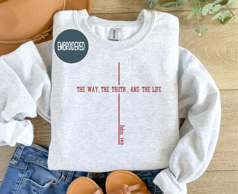 May include: Light gray embroidered sweatshirt with the text "THE WAY, THE TRUTH, AND THE LIFE" in red, with "John 14:6" below. The sweatshirt has a crew neck and long sleeves. A circular patch that says "EMBROIDERED" is on the upper left.