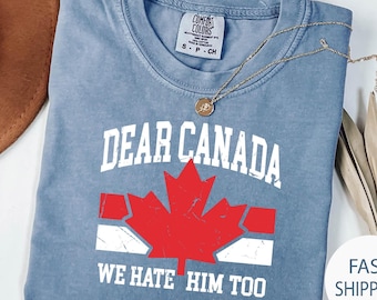 May include: A blue Comfort Colors t-shirt with the text "DEAR CANADA WE HATE HIM TOO" and a red maple leaf graphic. A gold necklace with a pendant is draped over the shirt.