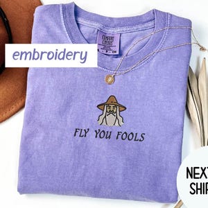 May include: A light purple t-shirt with an embroidered wizard design and the text "FLY YOU FOOLS". The shirt has a crew neck and a small tag. A gold necklace with a pendant is draped across the shirt.