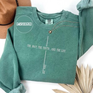 May include: A teal green sweatshirt with the text "THE WAY, THE TRUTH, AND THE LIFE" and "John 14:6" printed in white. The word "EMBROIDERED" is in a circle on the upper left. A gold necklace is draped across the front.