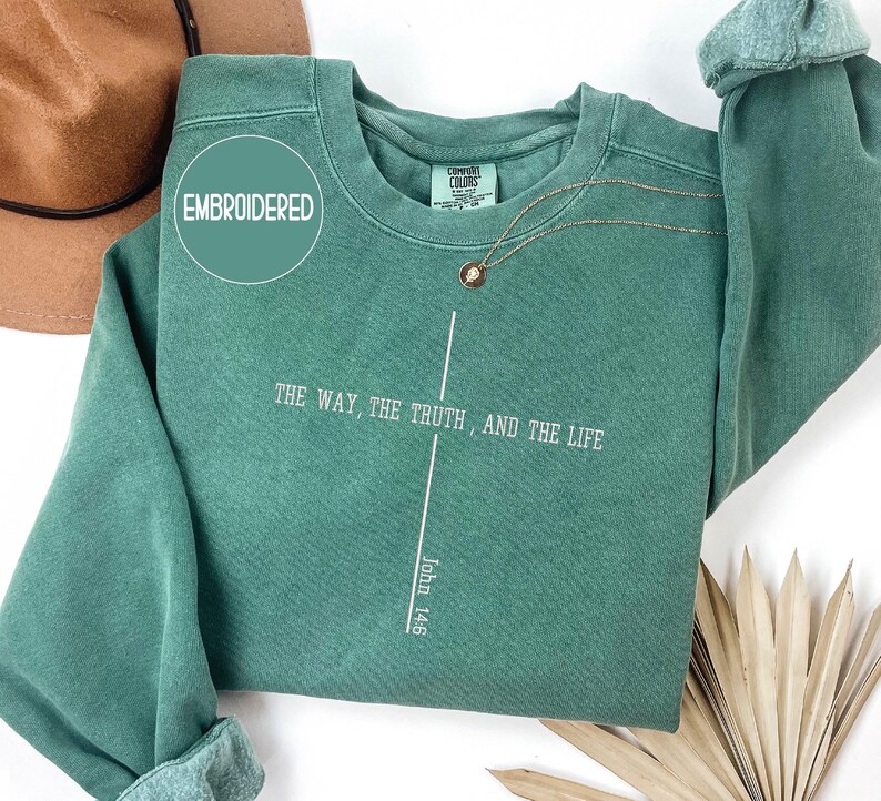 May include: A teal green sweatshirt with the text "THE WAY, THE TRUTH, AND THE LIFE" and "John 14:6" printed in white. The word "EMBROIDERED" is in a circle on the upper left. A gold necklace is draped across the front.