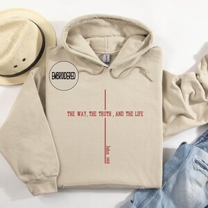 May include: A beige hoodie with a drawstring hood and a front pocket. The front of the hoodie features a red cross design with the text "THE WAY, THE TRUTH, AND THE LIFE" and "John 14:6". A straw hat is visible in the upper left corner.