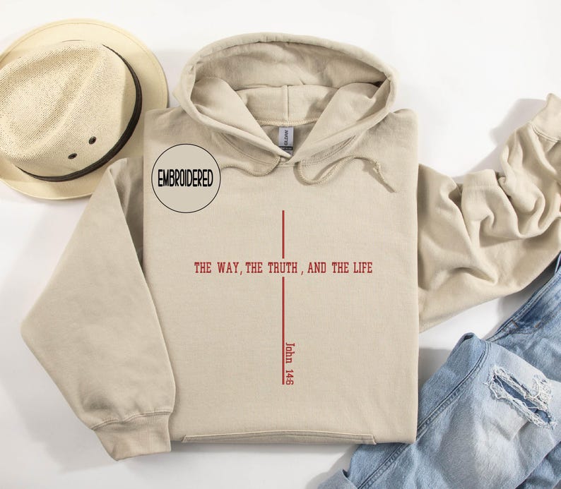 May include: A beige hoodie with a drawstring hood and a front pocket. The front of the hoodie features a red cross design with the text "THE WAY, THE TRUTH, AND THE LIFE" and "John 14:6". A straw hat is visible in the upper left corner.