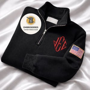 May include: Black quarter-zip sweatshirt with red embroidered monogram and an American flag patch on the sleeve. The sweatshirt features a zipper closure and ribbed cuffs. The image includes a circular logo that says "Embroidered Custom Thread Color."