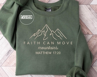 May include: An olive green sweatshirt with the embroidered word "EMBROIDERED" in a circle. The sweatshirt features a mountain graphic and the text "FAITH CAN MOVE mountains MATTHEW 17:20" in a light beige color.