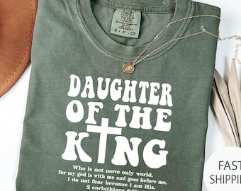 May include: A moss green t-shirt with the text "DAUGHTER OF THE KING" in white. The shirt also features a quote from 2 Corinthians 6:18. A gold necklace with a pendant rests on the shirt.