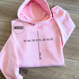 May include: A light pink hoodie with a matching hood lining. The front of the hoodie features the text "THE WAY, THE TRUTH, AND THE LIFE" with a vertical line and the text "John 14:6". The word "EMBROIDERED" is in a circle on the upper left.