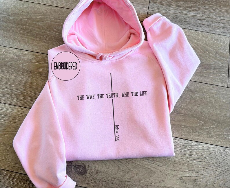 May include: A light pink hoodie with a matching hood lining. The front of the hoodie features the text "THE WAY, THE TRUTH, AND THE LIFE" with a vertical line and the text "John 14:6". The word "EMBROIDERED" is in a circle on the upper left.