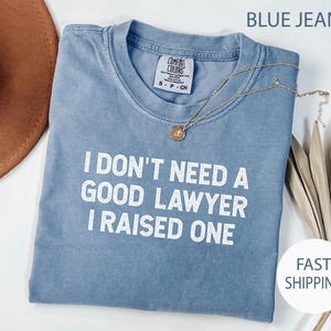 May include: A blue Comfort Colors t-shirt with the text "I DON'T NEED A GOOD LAWYER I RAISED ONE" in white. A gold necklace with a pendant is draped across the shirt. The shirt is folded.