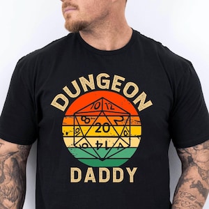 May include: Black t-shirt with a retro design. The graphic features a D20 die and the text "DUNGEON DADDY" in gold. The design incorporates orange, yellow, and green, appealing to fans of tabletop role-playing games.