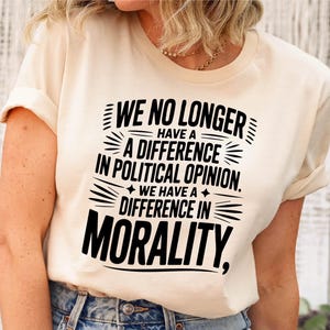 May include: Cream-colored t-shirt with black text. The text reads "WE NO LONGER HAVE A DIFFERENCE IN POLITICAL OPINION. WE HAVE A DIFFERENCE IN MORALITY." The shirt has short sleeves and a crew neck.