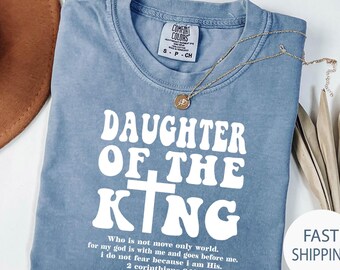 May include: A blue t-shirt with the text "DAUGHTER OF THE KING" in white. The shirt also features a quote from 2 Corinthians 6:18. A gold necklace with a pendant rests on the shirt. The shirt is made of a soft material.