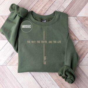 May include: An olive green sweatshirt with the text "THE WAY, THE TRUTH, AND THE LIFE" and "John 14:6" printed in a vertical line. The word "EMBROIDERED" is in a white circle. The sweatshirt is folded on a light wood surface.