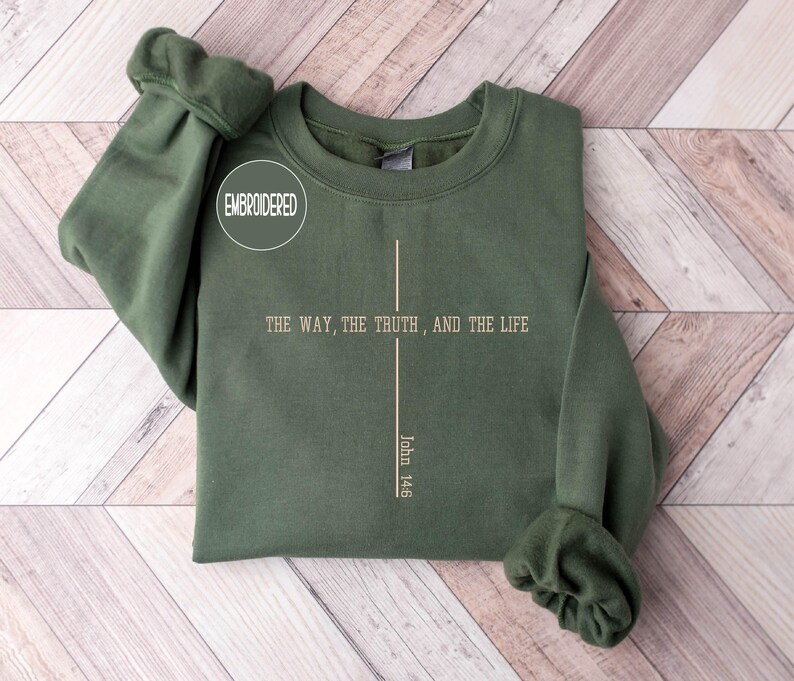 May include: An olive green sweatshirt with the text "THE WAY, THE TRUTH, AND THE LIFE" and "John 14:6" printed in a vertical line. The word "EMBROIDERED" is in a white circle. The sweatshirt is folded on a light wood surface.