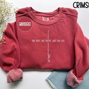 May include: Crimson-colored embroidered sweatshirt with the text "THE WAY, THE TRUTH, AND THE LIFE" and "John 14:6" in white. The sweatshirt has a crew neck and cuffed sleeves. The word "EMBROIDERED" is in a circle.