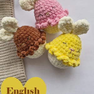 Puede incluir: Tres juguetes de conejito de ganchillo hechos a mano en rosa, marrón y amarillo. Cada conejito tiene orejas color crema y un borde con volantes. Los juguetes están sobre una superficie blanca con un artículo de punto beige y hojas secas en el fondo. El texto "English Pattern" está en un corazón amarillo.