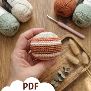 May include: A crocheted ball in shades of brown, orange, white, and blue is held in a hand. The ball is surrounded by yarn, a crochet hook, and decorative elements on a wooden surface. The image includes the text "PDF PATTERN".