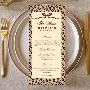 Peut inclure: Un menu pour l'anniversaire d'Olivia, avec un motif léopard. Le menu comprend des entrées, des plats principaux et des desserts. La carte est posée sur une assiette blanche avec une fourchette et un couteau dorés.
