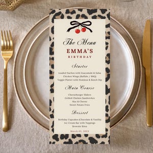 May include: A menu card for Emma's birthday, featuring a leopard print border. The menu includes starters, main courses, and desserts. The card is placed on a plate with a gold fork and knife.