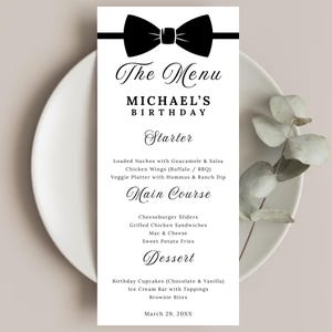 Könnte beinhalten: Eine Menükarte mit einer schwarzen Fliege oben. Das Menü für Michaels Geburtstag listet Vorspeisen, Hauptgerichte und Desserts auf. Das Datum ist der 29. März 20XX. Die Karte liegt auf einem weißen Teller.