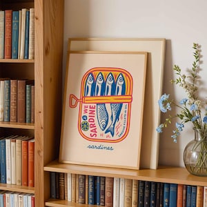 May include: A framed art print featuring a colorful illustration of a sardine can. The artwork depicts sardines in oil with blue, yellow, and red accents. The print is displayed on a wooden shelf with a bookcase and a vase of blue flowers.