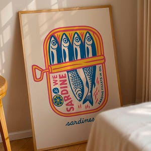 May include: A framed art print featuring a colorful illustration of a sardine can. The can is yellow and pink, with blue sardines inside. Text on the can reads "WE SARDINE" and "SARDINE FILLETS IN OIL". The word "sardines" is written at the bottom.