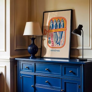 May include: A blue dresser with brass knobs and a framed poster featuring a sardine can illustration. The poster has the words "WE SARDINE" and "sardines" printed on it. A navy blue lamp and a black desk lamp are on top of the dresser.