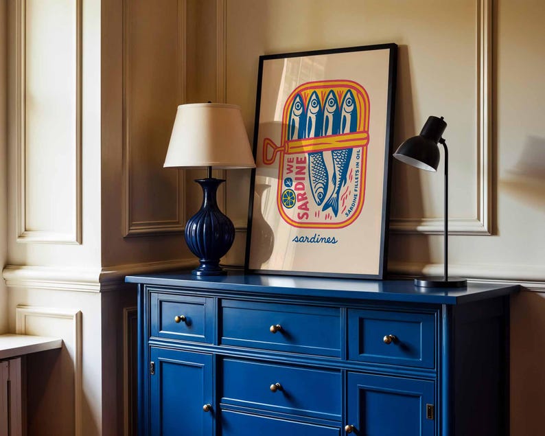 May include: A blue dresser with brass knobs and a framed poster featuring a sardine can illustration. The poster has the words "WE SARDINE" and "sardines" printed on it. A navy blue lamp and a black desk lamp are on top of the dresser.