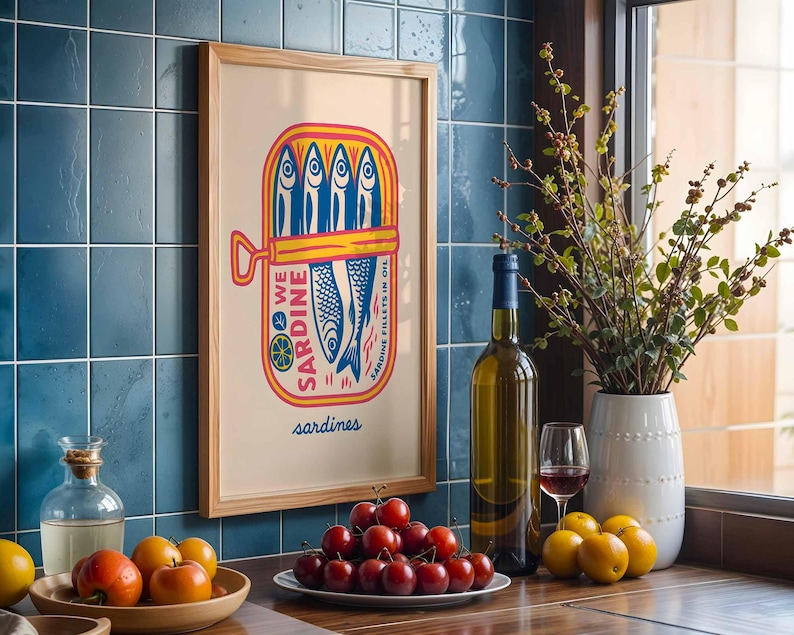 May include: A framed print featuring a colorful sardine can illustration with the text "WE SARDINE" and "SARDINE FILLETS IN OIL". The image is set in a kitchen with a bottle of wine, a glass of red wine, and a vase of flowers.