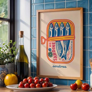 May include: A framed print featuring a colorful illustration of sardines in a can. The artwork includes the words "WE SARDINE" and "SARDINE FILLETS IN OIL". The print is displayed near a window, a bottle of olive oil, and fresh produce.