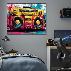 Może przedstawiać: Kolorowy obrazek przedstawiający zabytkowy boombox. Grafika przedstawia żółto-różowy zestaw stereo z niebieskimi i pomarańczowymi akcentami. Obraz jest oprawiony i wisi na szarej ścianie nad łóżkiem i biurkiem.