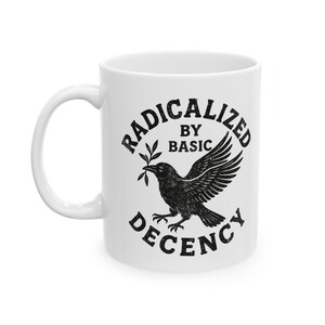 May include: White ceramic mug with the words "RADICALIZED BY BASIC DECENCY" in a black, distressed font. A black illustration of a crow holding a sprig in its beak is centered on the mug. The mug has a white handle.