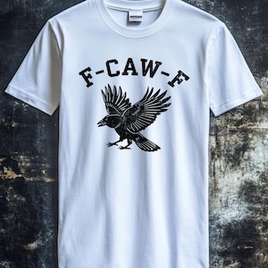 Puede incluir: Camiseta blanca con un gráfico negro de un cuervo con las alas extendidas y el texto "F-CAW-F" encima. El diseño está centrado en la camiseta, que tiene cuello redondo y mangas cortas. El fondo es una superficie oscura y texturizada.