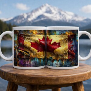 Könnte beinhalten: Zwei weiße Keramikbecher mit einem kanadischen Flaggen-Design. Die Flagge zeigt ein rotes Ahornblatt auf weißem Grund, vor einem strukturierten, künstlerischen Hintergrund mit Gold-, Blau- und Gelbtönen. Die Becher stehen auf einem Holzschemel.