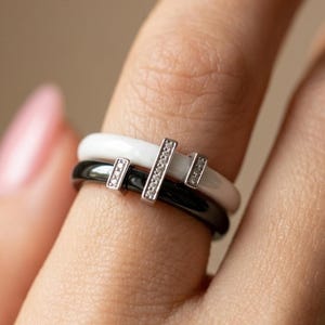 Peut inclure: Deux bagues minimalistes, une blanche et une noire, superposées sur un doigt. Chaque bague est ornée d'une barre argentée avec de petits ornements scintillants. Ces bagues sont un design de bijoux moderne et élégant.