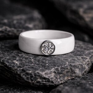May include: A white ceramic ring with a round, silver-toned setting. The setting is encrusted with small, clear gemstones. The ring is displayed on a dark gray, textured stone surface.