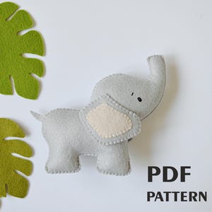 May include: A grey felt elephant toy with a beige felt ear. The toy is standing on a white surface with green felt leaves in the background. The text "PDF PATTERN" is printed in black below the toy.
