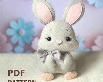 Può includere: Un coniglietto di feltro grigio con orecchie rosa e un fiocco grigio. Il coniglietto ha occhi neri e un naso rosa. Il coniglietto è seduto su una superficie bianca. MODELLO PDF.
