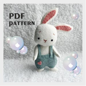 May include: A handmade white felt bunny wearing blue overalls with a pink heart. The bunny has pink inner ears and black eyes. The image includes the text "PDF PATTERN" in black letters and decorative bubbles.
