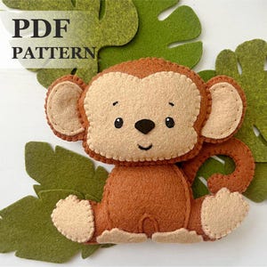 Può includere: Un modello di giocattolo di scimmia in feltro. La scimmia è marrone con un viso beige e ha un sorriso cucito. Il modello è per un download PDF.