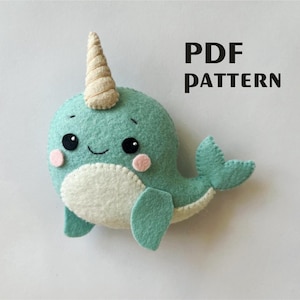 May include: A handmade teal felt narwhal plushie with a cream-colored horn. The narwhal has a white belly, pink cheeks, and black eyes. The text "PDF PATTERN" is printed in black.