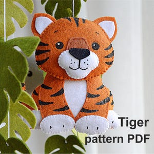 May include: A felt tiger toy with orange and black stripes, white paws, and a white belly. The toy is hanging from a string and has a white face with black eyes and a black nose. The text "Tiger pattern PDF" is visible below the toy.