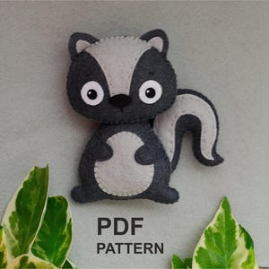 Puede incluir: Un mapache de fieltro gris y negro con la nariz y los ojos blancos. El mapache está de pie sobre un fondo gris con hojas verdes a cada lado. El texto "PDF PATTERN" está impreso debajo del mapache.