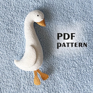 May include: A white felt goose toy with a long neck, small black eyes, and a tan beak and feet. The goose is on a light blue textured surface. Text on the right reads "PDF PATTERN".