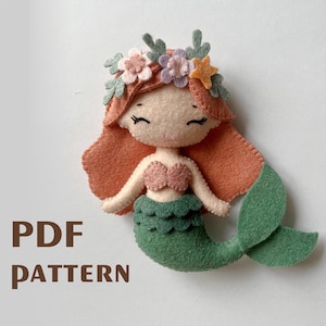 Puede incluir: Una muñeca de sirena de fieltro con cola verde, cabello naranja y una corona de flores. La muñeca tiene un sujetador de concha rosa y una cara sonriente.